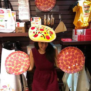 Woman holding up two fake pizzas wearing a pizza hat in front of a background of pizza souvenirs (They probably don't like pizza in Chicago)