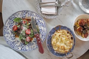 Spread of steak, macaroni and cheese and scallops on different patterned blue and white plates on marble table at The Allis Restaurant in the Soho house Chicago